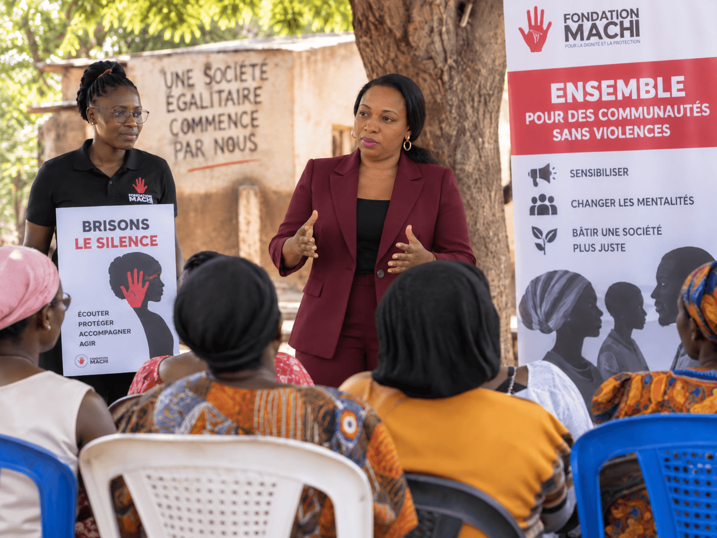Une intervenante de la Fondation s’adresse à un groupe de femmes assises en plein air, devant des affiches de campagne.